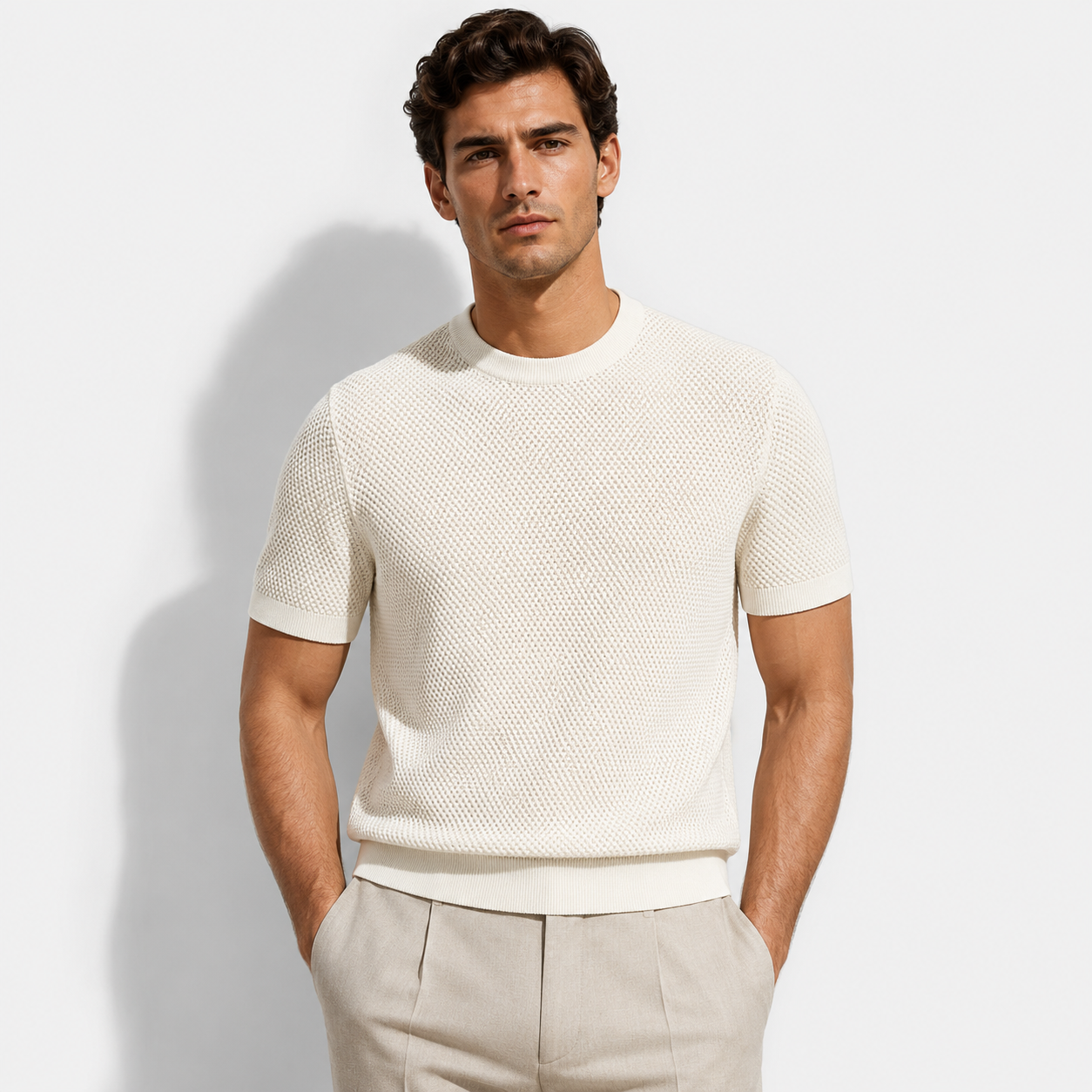 A man wearing a short-sleeve textured knit crewneck sweater in off-white, paired with light beige trousers, standing against a light gray studio background.