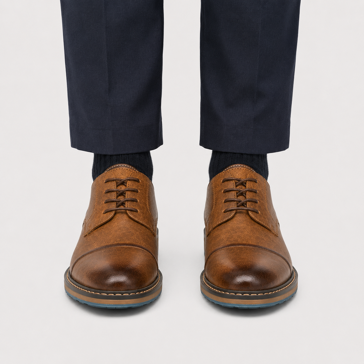A pair of men's brown leather lace-up dress shoes with a cap toe, shown on a model wearing navy trousers and dark socks against a light gray background.