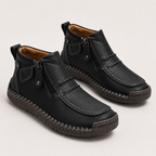 A pair of black leather ankle boots with side zippers, moc-toe stitching, and a braided welt, shown on a light gray background.