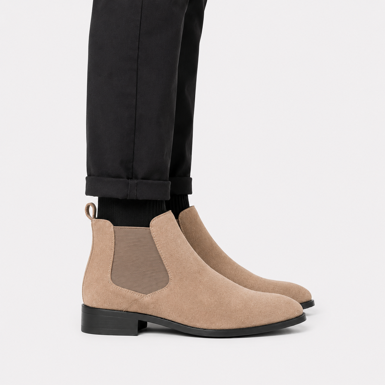 A pair of beige suede Chelsea boots with black soles and black elastic side panels, shown on a model wearing black trousers against a light gray background.