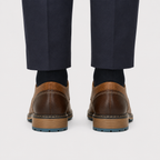 Rear view of a person wearing dark navy trousers, black socks, and brown leather shoes with a tan midsole and blue outsole, shown against a light gray studio background.