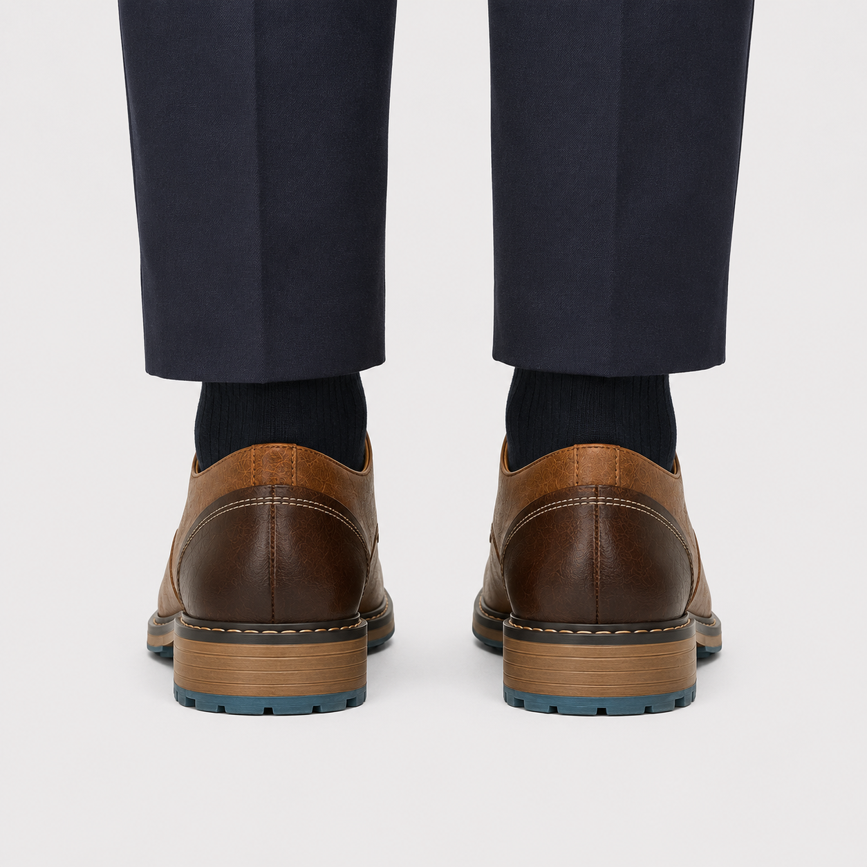 Rear view of a person wearing dark navy trousers, black socks, and brown leather shoes with a tan midsole and blue outsole, shown against a light gray studio background.