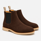 A pair of dark brown suede Chelsea boots with black elastic side panels and light tan pull tabs and soles, shown on a clean white background.