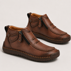 A pair of brown faux-leather ankle boots with side zippers, decorative stitching, and pull tabs, shown on a light gray background.