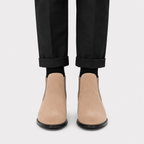 A pair of beige suede Chelsea-style ankle boots worn with black socks and black trousers, shown from the front against a light gray background.