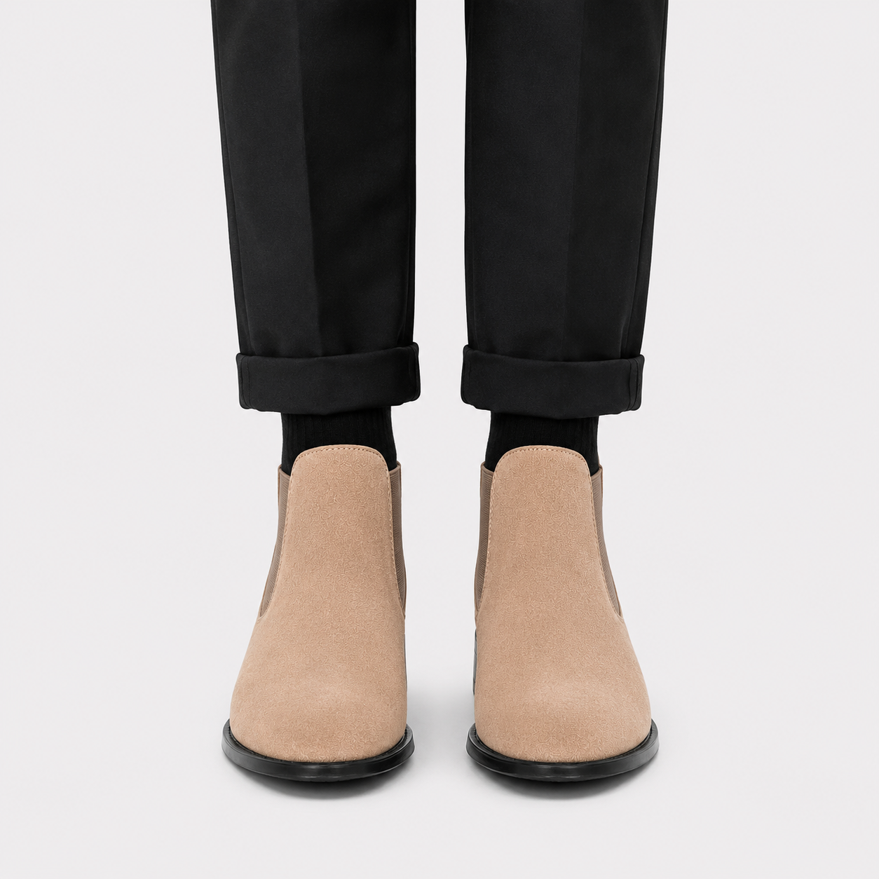 A pair of beige suede Chelsea-style ankle boots worn with black socks and black trousers, shown from the front against a light gray background.