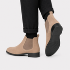 A pair of beige suede Chelsea-style ankle boots with black soles, shown on a model wearing black cuffed trousers against a light gray studio background.