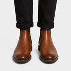 A pair of men's brown leather Chelsea boots worn with black cuffed trousers, shown from the front against a light gray background.