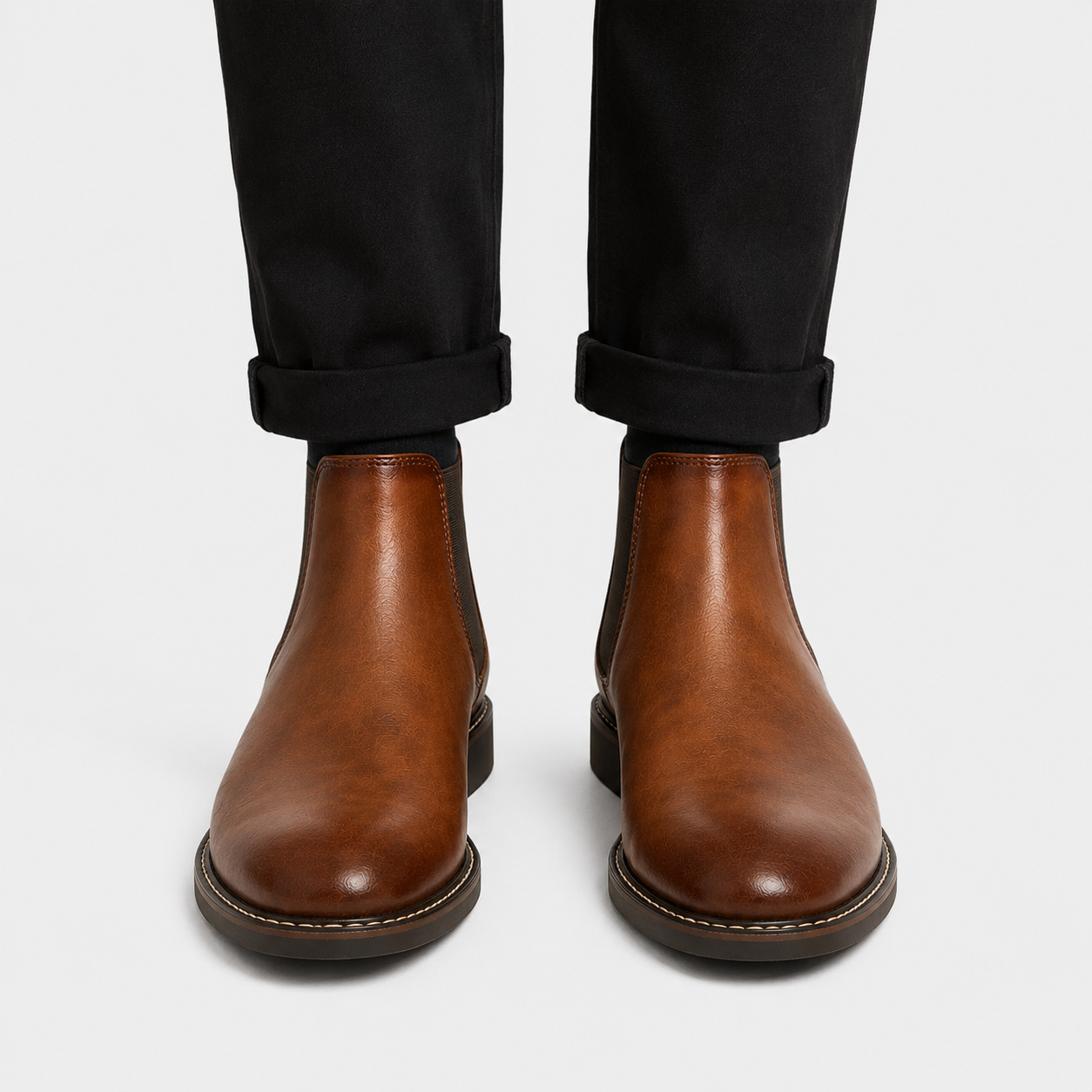 A pair of men's brown leather Chelsea boots worn with black cuffed trousers, shown from the front against a light gray background.