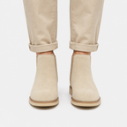 A pair of beige suede Chelsea-style ankle boots worn with light beige rolled-up trousers, shown from the front against a light gray studio background.