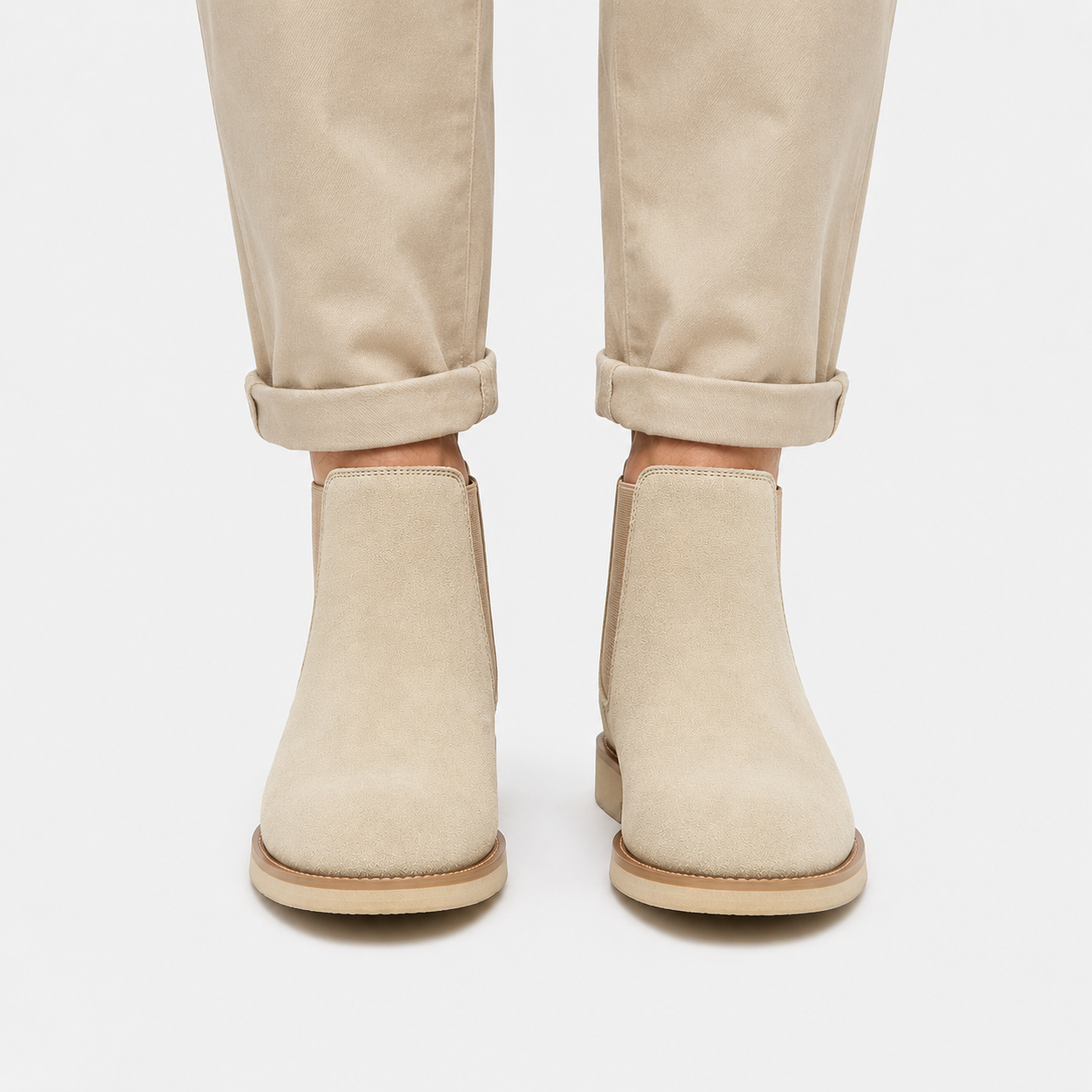A pair of beige suede Chelsea-style ankle boots worn with light beige rolled-up trousers, shown from the front against a light gray studio background.