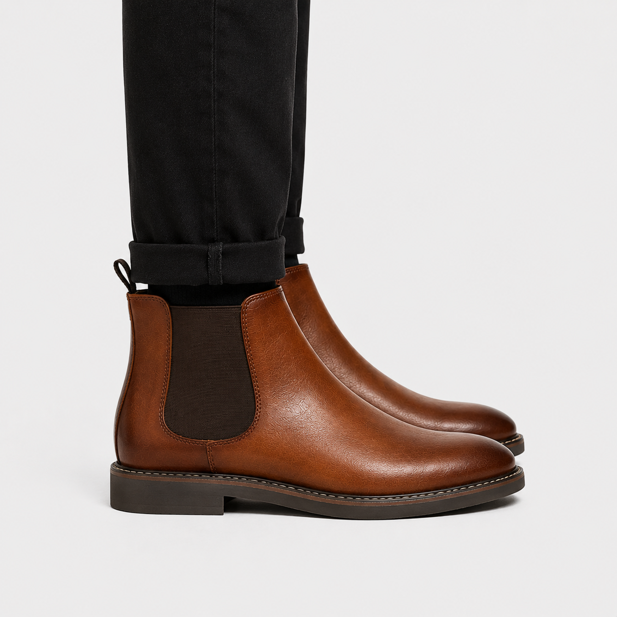 A pair of men's brown leather Chelsea boots with dark elastic side panels, shown worn with black cuffed trousers against a light gray background.