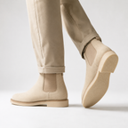 Beige suede Chelsea-style ankle boots worn with light beige trousers, shown from the side and back on a clean light gray studio background.