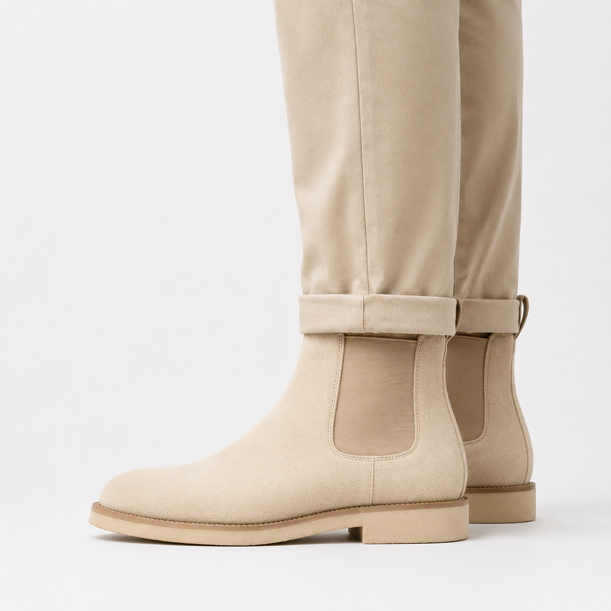 Beige suede Chelsea-style ankle boots with elastic side panels, shown on a model wearing matching beige trousers against a clean white background.
