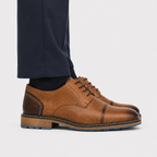 A pair of men's brown leather oxford shoes with dark brown paneling and laces, styled with dark navy trousers and black socks on a light gray background.