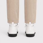 White ankle boots with black soles and pull tabs, shown from the back on a model wearing beige pants against a light gray background.