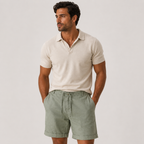 A man wearing a light beige short-sleeve polo shirt and sage green drawstring shorts, standing against a plain light gray studio background.