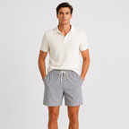 A man modeling a cream short-sleeve polo shirt and gray-and-white vertically striped drawstring shorts against a light gray studio background.