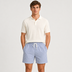 A man modeling a short-sleeve polo shirt in off-white and blue-and-white vertically striped shorts against a light gray studio background.