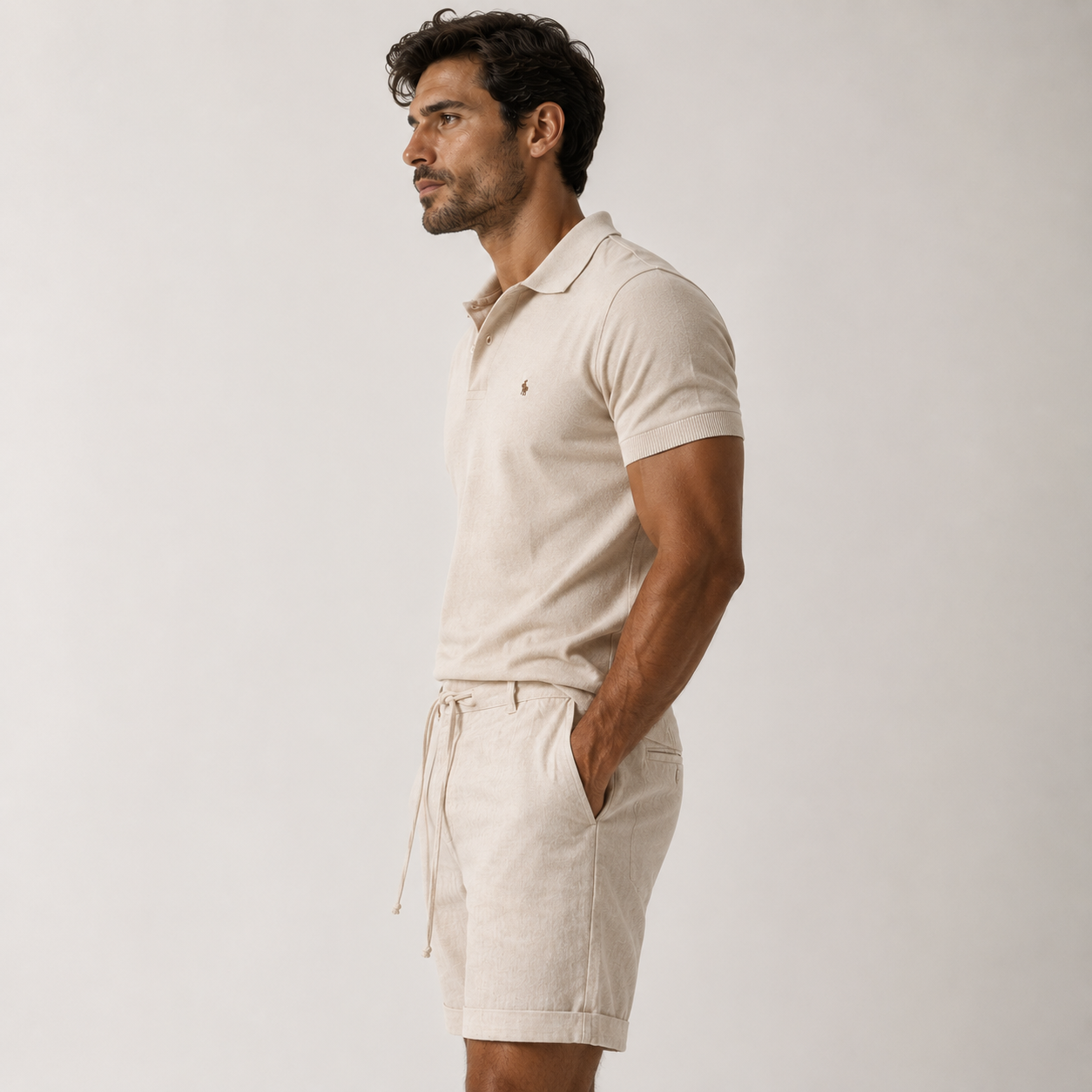 A man in side profile wearing a light beige short-sleeve polo shirt and matching drawstring shorts against a plain light gray studio background.