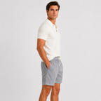 A man wearing a white short-sleeve polo shirt and gray and white vertically striped drawstring shorts, standing in profile against a light gray studio background.
