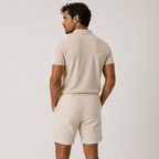 A man shown from the back wearing a light beige short-sleeve polo shirt and matching light beige shorts against a plain light gray studio background.
