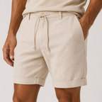 Beige linen drawstring shorts worn by a model, featuring a relaxed fit and rolled cuffs, paired with a matching beige top against a light gray background.