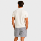 A man viewed from behind wearing a cream short-sleeve polo shirt and gray and white striped shorts, set against a solid light gray background.