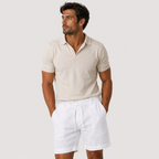 A man wearing a light beige short-sleeve polo shirt and white drawstring shorts, standing against a light gray studio background.