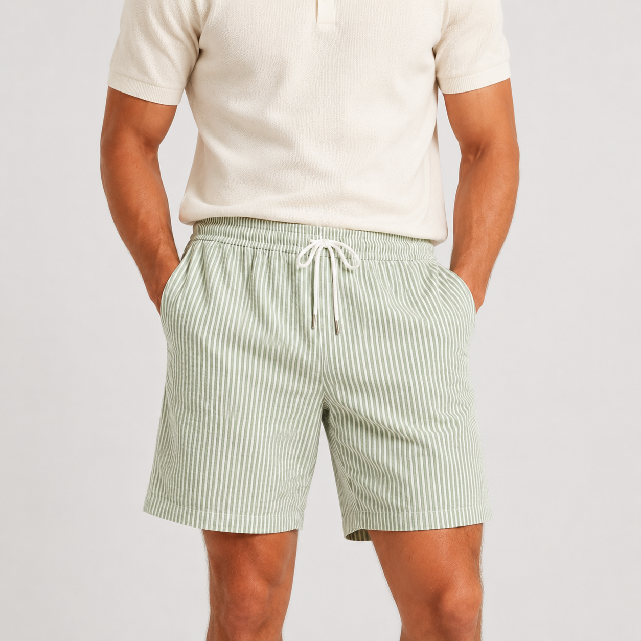 A man wearing light green and white vertically striped drawstring shorts with a cream polo shirt, shown from the chest down against a light gray studio background.