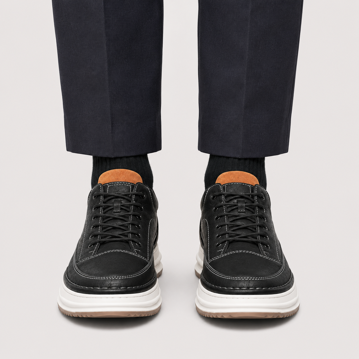 Black leather low-top lace-up sneakers with thick white platform soles, shown on a model wearing dark trousers and black socks against a light gray background.