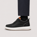 A black leather low-top sneaker with a thick white platform sole and a brown heel tab, shown on a model wearing dark trousers and black socks against a light gray background.