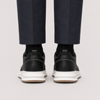 Rear view of a person wearing black leather low-top sneakers with chunky white midsoles and dark navy trousers, shown against a light gray studio background.
