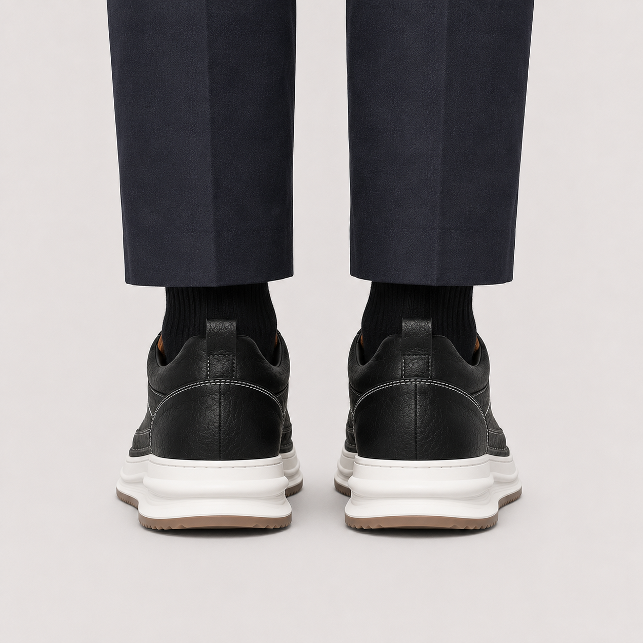 Rear view of a person wearing black leather low-top sneakers with chunky white midsoles and dark navy trousers, shown against a light gray studio background.