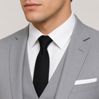 A close-up of a man wearing a light gray three-piece suit, white dress shirt, and black tie against a plain light gray background.