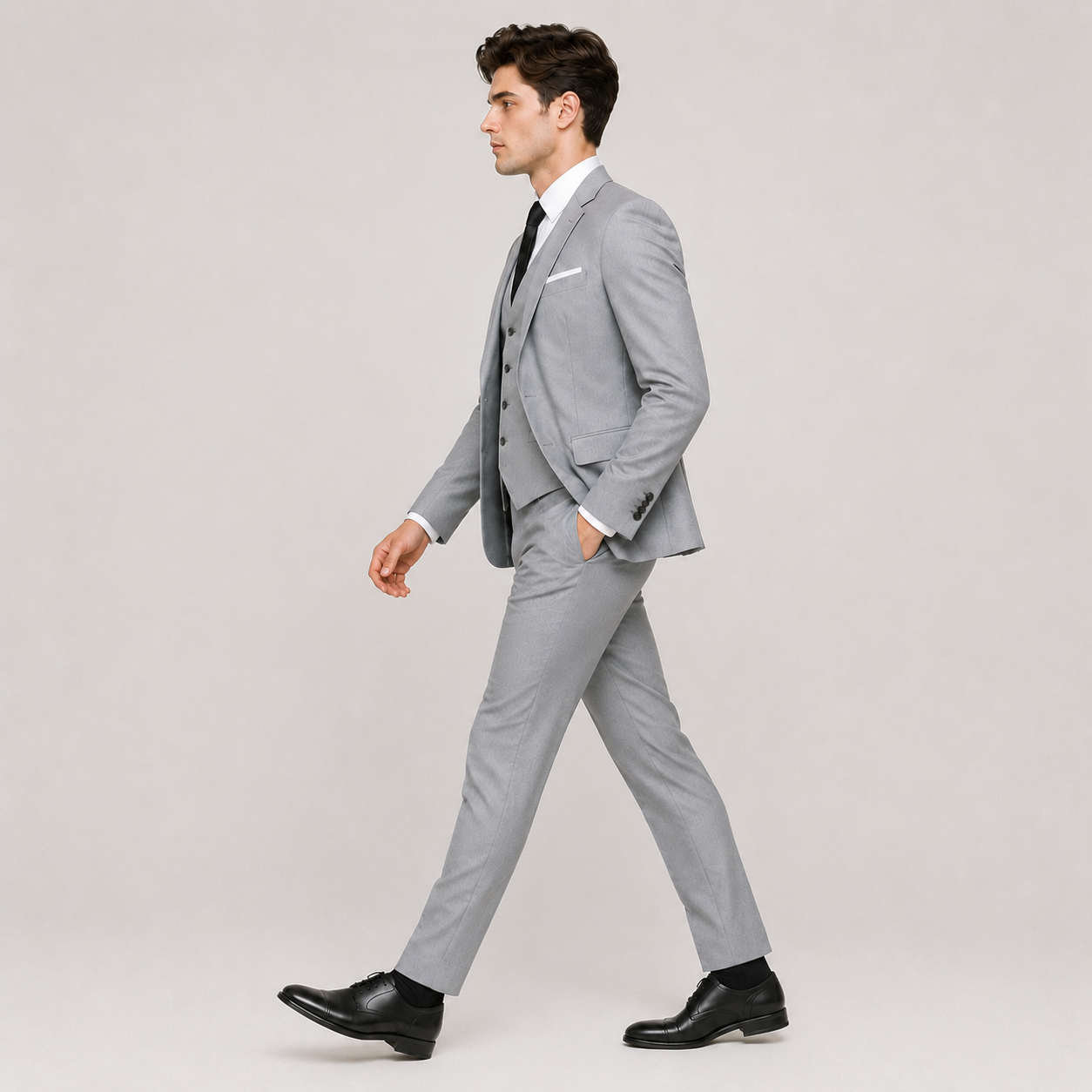 A man wearing a light gray three-piece suit, white dress shirt, black tie, and black dress shoes, walking in profile against a plain light gray studio background.