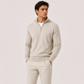 A man wearing a light beige quarter-zip knit sweater over a white undershirt and matching beige trousers, standing against a plain light beige studio background.