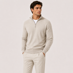 A man wearing a light beige quarter-zip knit sweater over a white undershirt and matching beige trousers, standing against a plain light beige studio background.