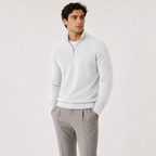 A man wearing a light gray quarter-zip knit sweater over a white undershirt and gray trousers, standing against a plain light gray studio background.
