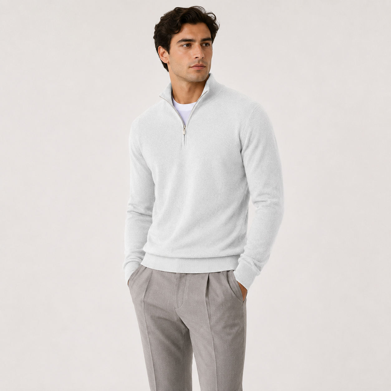 A man wearing a light gray quarter-zip knit sweater over a white undershirt and gray trousers, standing against a plain light gray studio background.