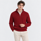 A man wearing a red cable-knit half-zip sweater over a white collared shirt and cream trousers, standing against a plain light gray studio background.