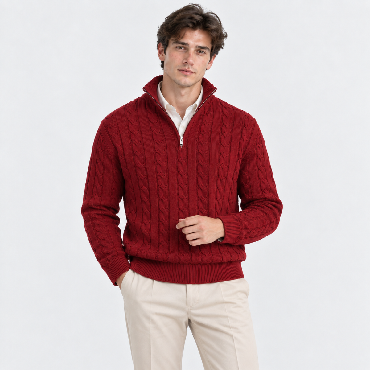 A man wearing a red cable-knit half-zip sweater over a white collared shirt and cream trousers, standing against a plain light gray studio background.