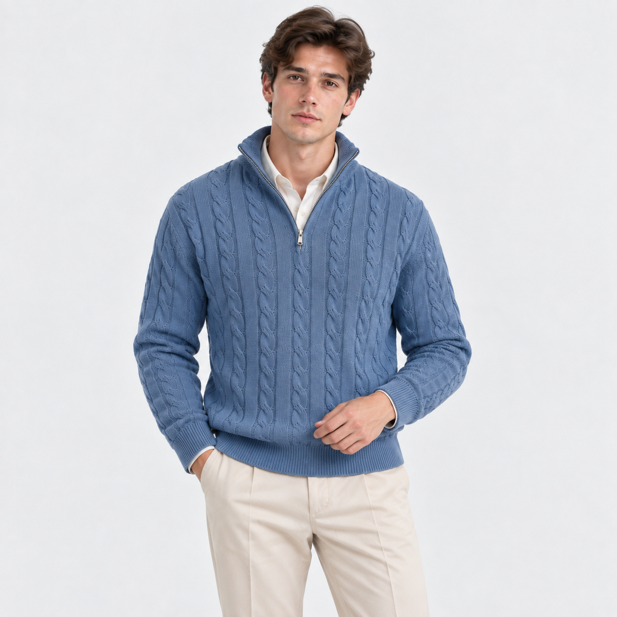 A man wearing a blue cable-knit quarter-zip sweater over a cream collared shirt and beige trousers, standing against a plain light gray studio background.