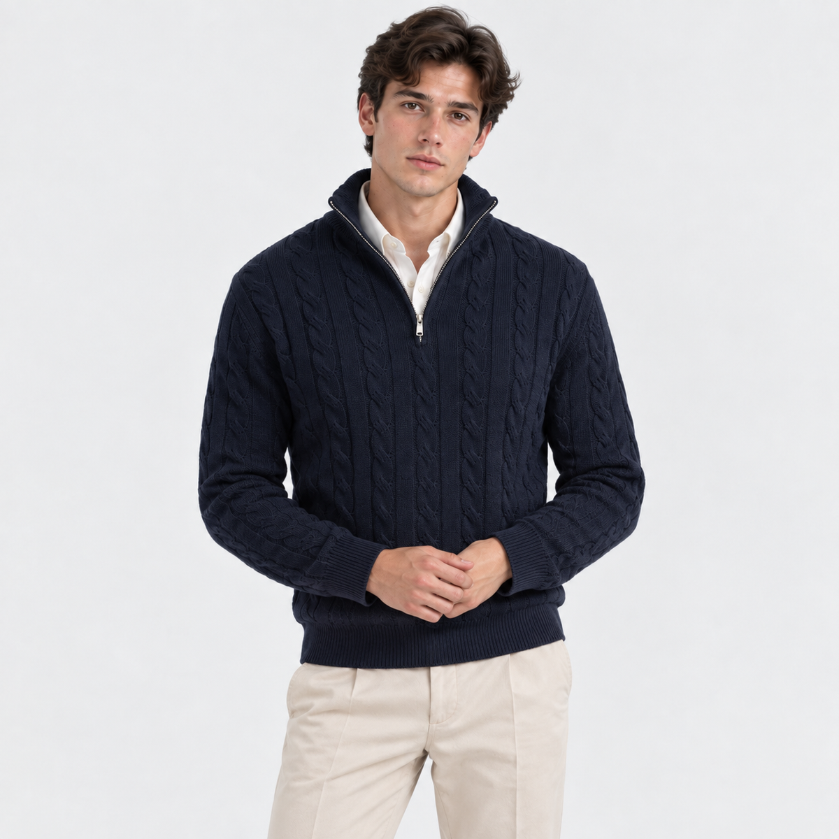 A man wearing a navy blue cable-knit quarter-zip sweater over a white collared shirt and beige trousers, standing against a plain white studio background.