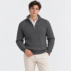 A man wearing a dark gray cable-knit quarter-zip sweater over a cream collared shirt and beige trousers, standing against a light gray studio background.