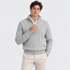 A man wearing a light gray cable-knit quarter-zip sweater over a white collared shirt and beige trousers, standing against a plain light gray studio background.