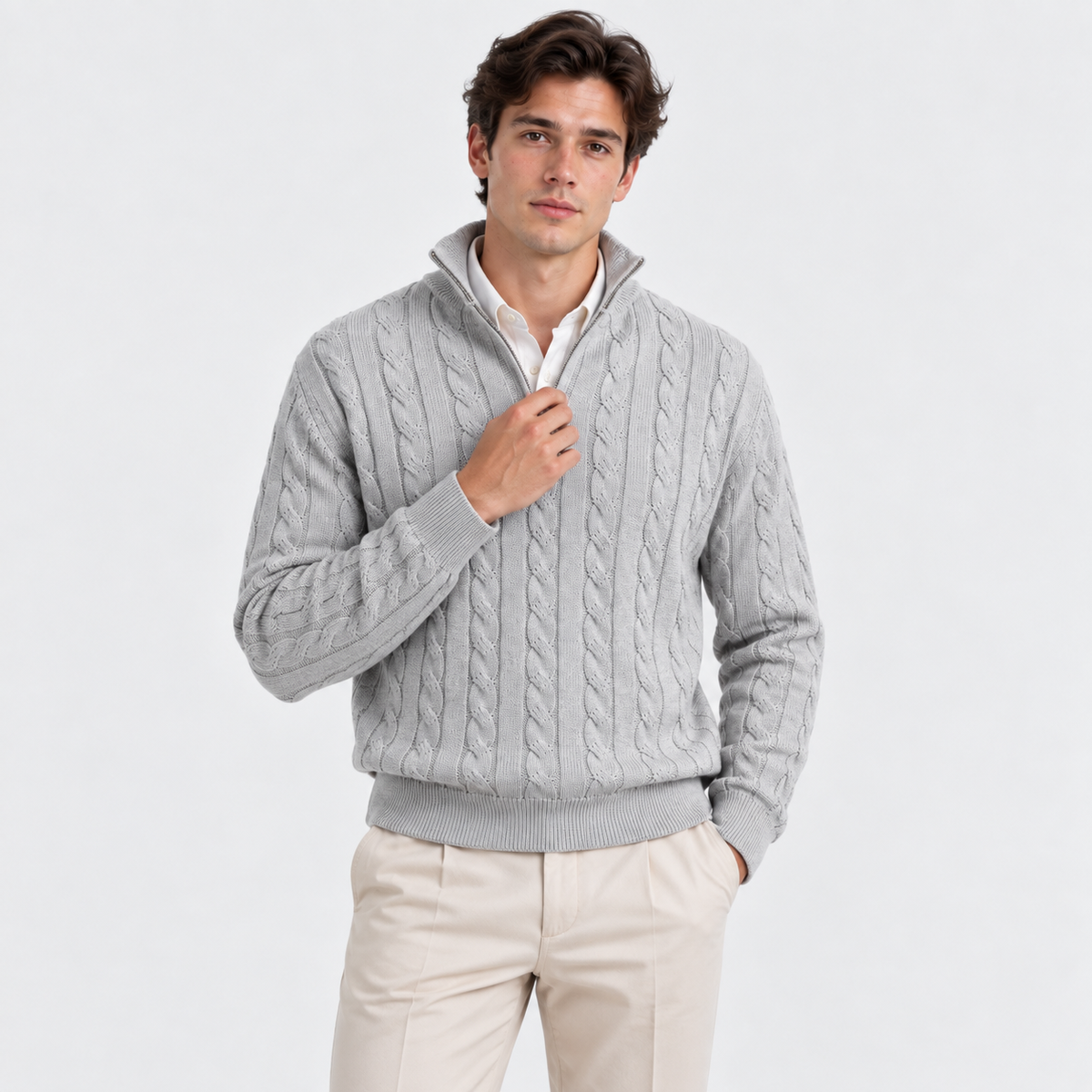 A man wearing a light gray cable-knit quarter-zip sweater over a white collared shirt and beige trousers, standing against a plain light gray studio background.