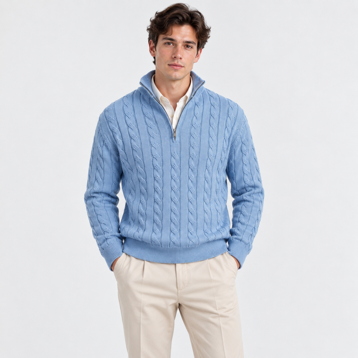 A man wearing a light blue cable-knit quarter-zip sweater over a cream collared shirt and beige trousers, standing against a plain light gray studio background.