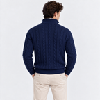 A man viewed from the back wearing a navy blue cable-knit sweater and light beige trousers, standing against a plain light gray studio background.