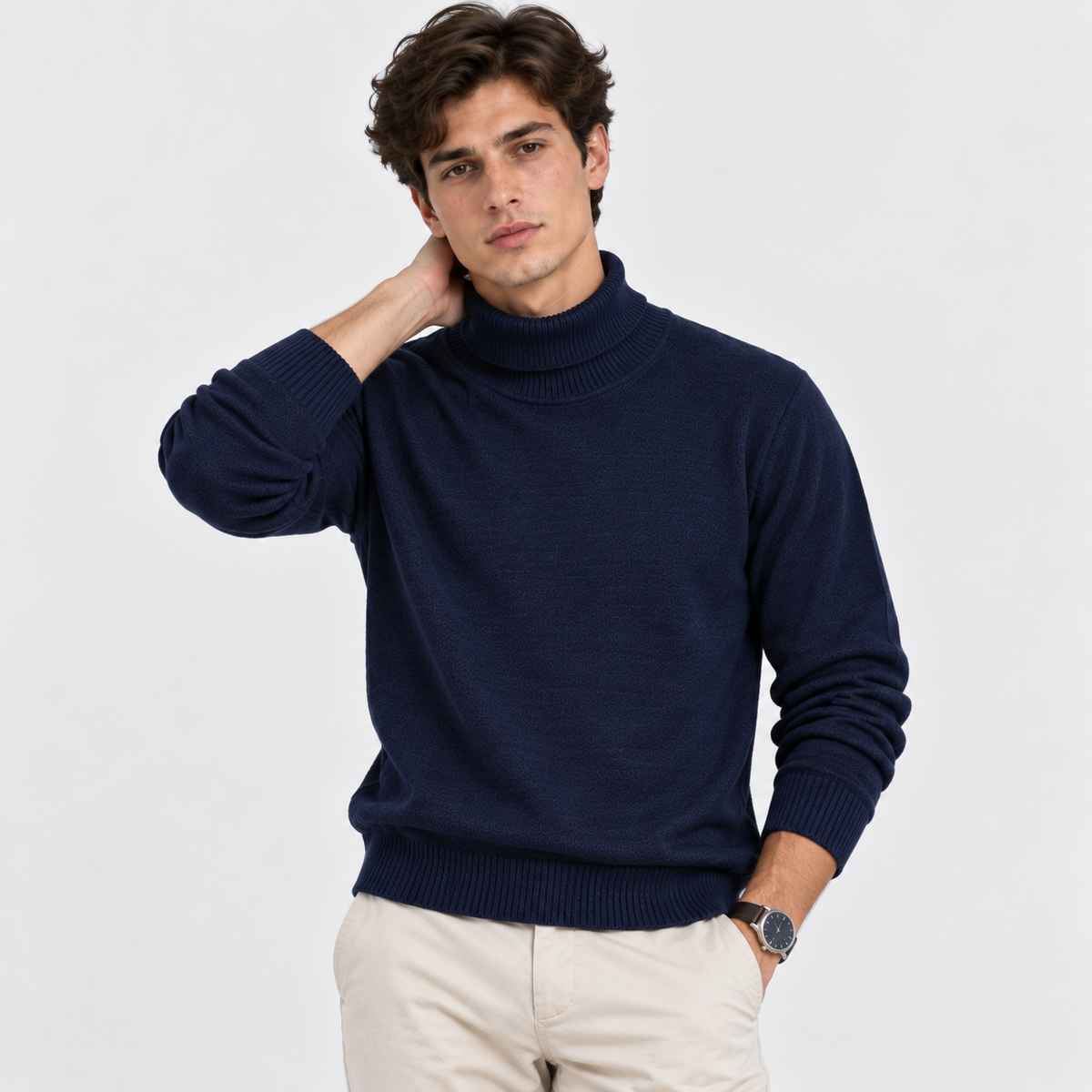 A man wearing a navy blue ribbed turtleneck sweater and beige trousers, posing against a light gray studio background.